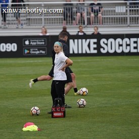 A person in sports attire stands on a soccer field, surrounded by several soccer balls and a carrier with bottles. The field is bordered by advertisements for Nike Soccer and app stores. Several people sit in bleachers in the background.