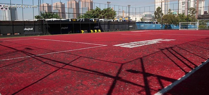 A sports court with a red playing surface and white lines, surrounded by a black mesh fence. In the background, tall buildings are visible, along with some trees and a few yellow chairs positioned near the fence.